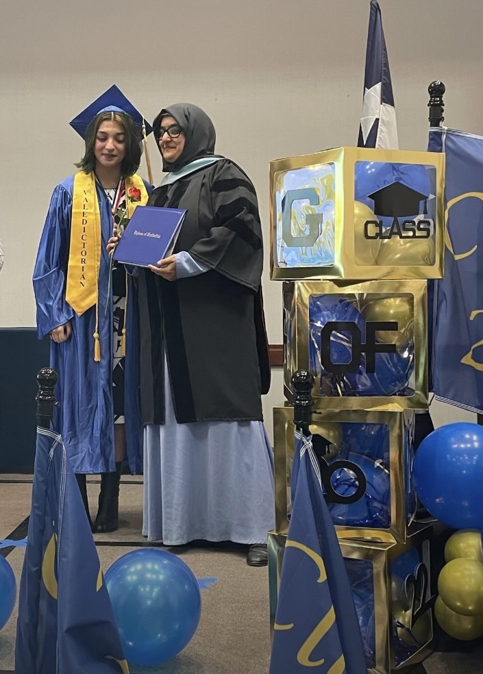 A hijabi principal hands a diploma to a high school graduate, dressed in royal blue graduation robes with a golden yellow sash